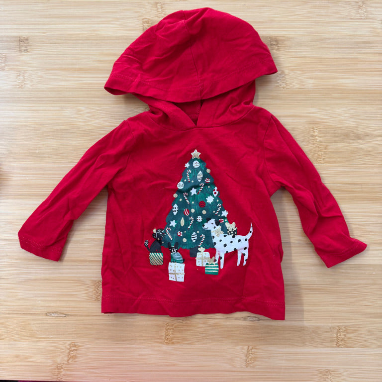 A red long-sleeve hooded baby/toddler top laid flat on a light wood surface, featuring a Christmas tree and presents graphic on the front with a white dog illustration on the right side of the tree.