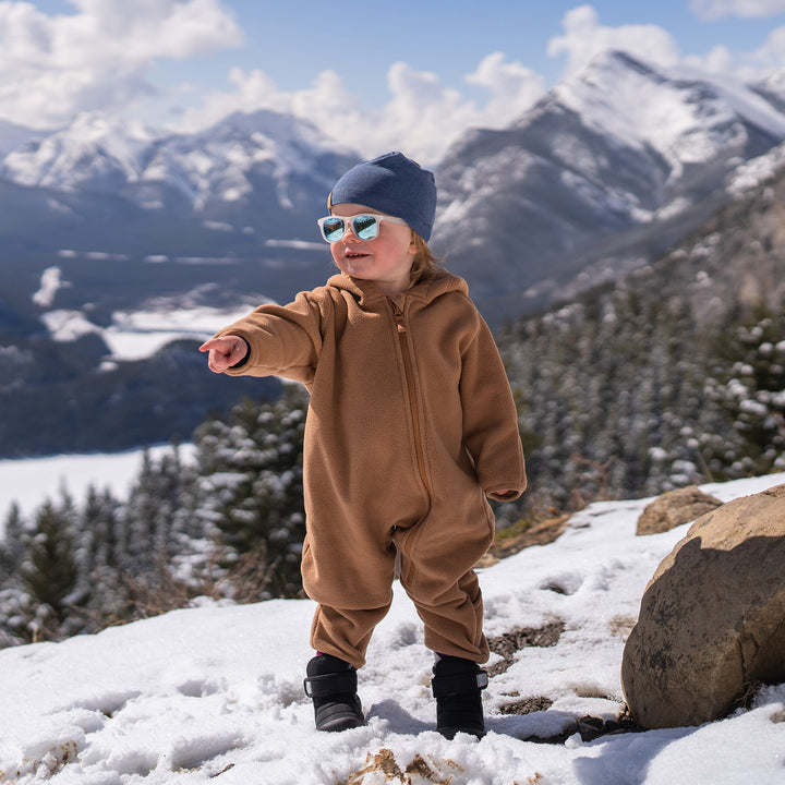 Child standing in the snow wearing the Jan & Jul Fleece Suit | Baby Outerwear in brown, wearing sunglasses, with mountains behind him and pointing at something 