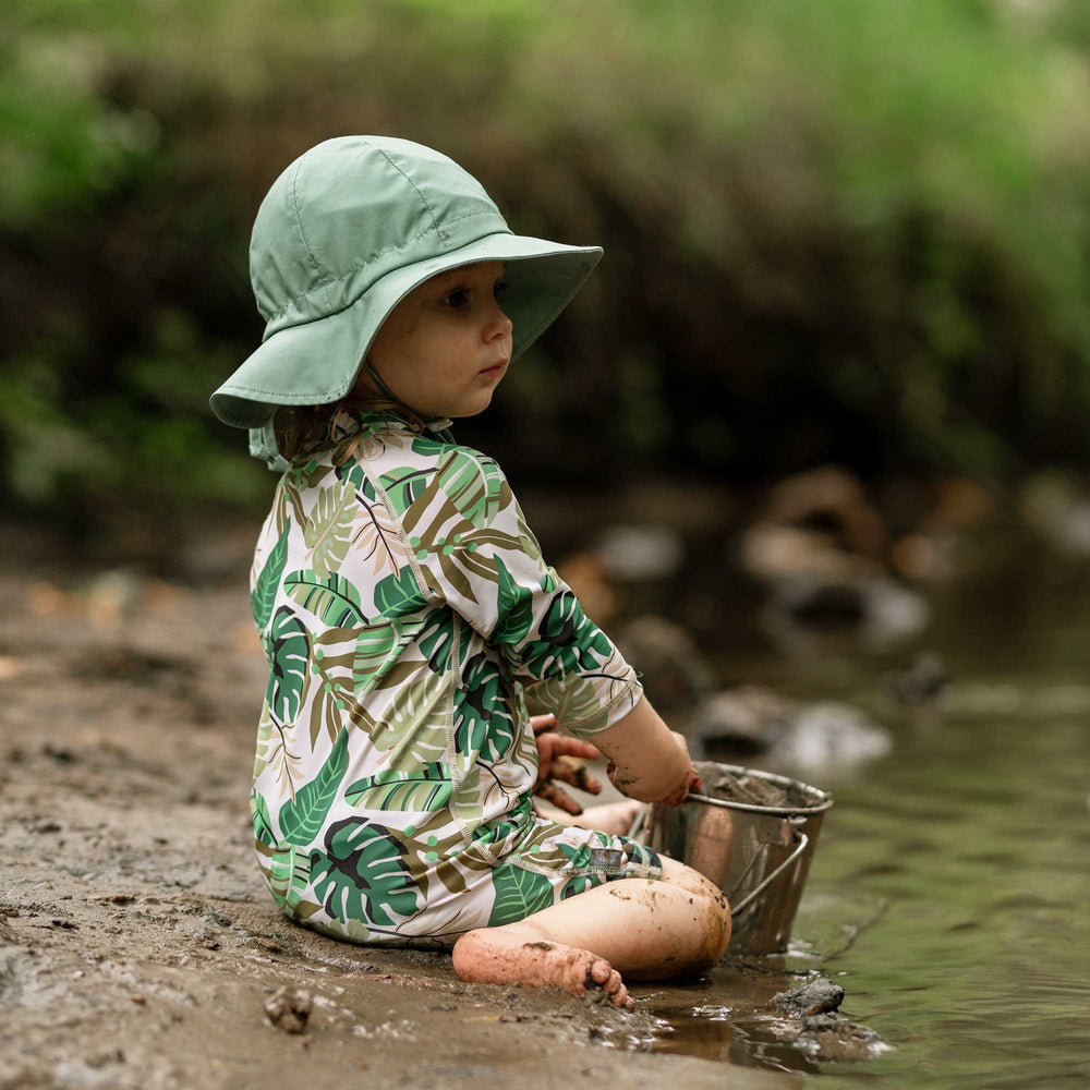 Child wearing th4e Jan & Jul Cotton Floppy Hat with coordinating sunsuit from Jan & Jul in Tropic Green in the mud at the beach