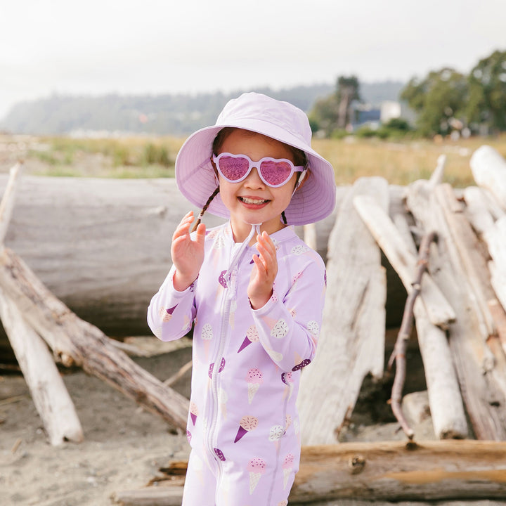 older child wearing Jan & Jul UV Jumpsuit | Lavendar Ice Cream with a coordinating bucket hat and sunglasses from Jan & Jul while at the beac