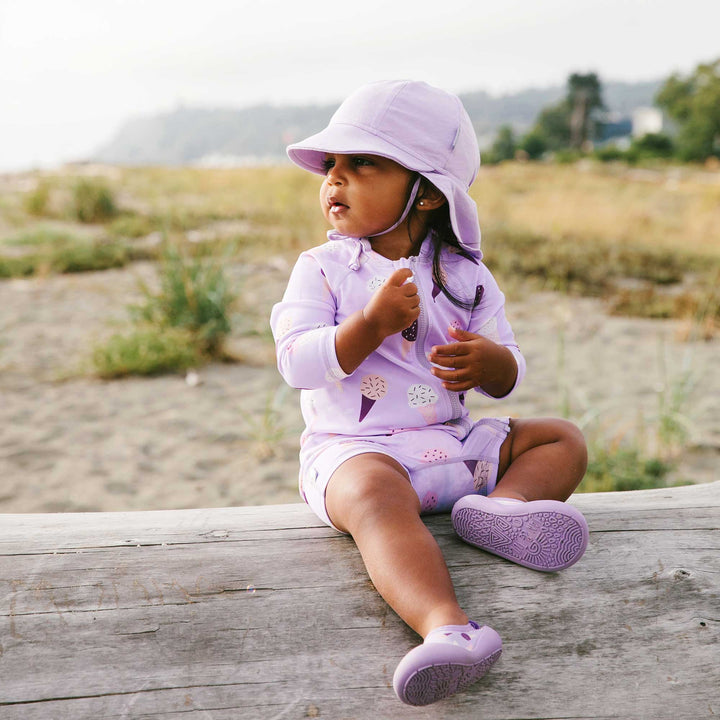 Child wearing the Jan & Jul UV Jumpsuit | Lavendar Ice Cream with a baby so soft cap and matching water play shoes on the beach 