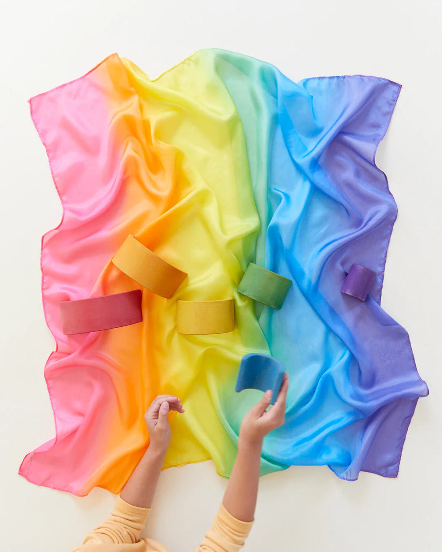 Rainbow Play Silk laid flat with wooden rainbow and childs hands playing on white back drop