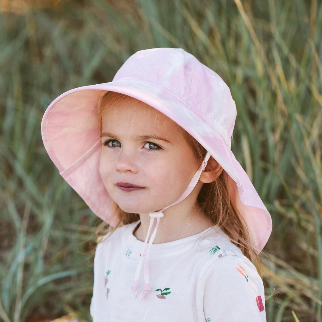 Jan & Jul Cotton Adventure Hat child wearing the pink ti dye variation in a field with full head coverage adn wide brim